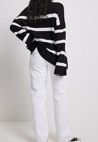 Schwarz-weißer gestreifter oversized Strickpullover mit weiten Ärmeln, kombiniert mit hochgeschnittenen weißen Jeans und schwarzen Sneakern.