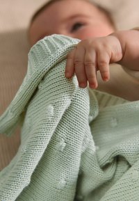 Couverture en tricot vert clair avec un motif texturé et des bords côtelés, tenue par une petite main, sur un fond neutre.