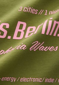 Groene stof met opvallende roze tekst, waaronder "3 steden // 1 nacht" en "Berlijn", met een gestructureerd patroon. Focus op typografische details.