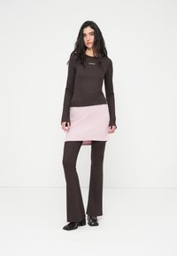 Καφέ μπλούζα με μακριά μανίκια και ανάγλυφη υφή με λογότυπο, συνδυασμένη με ροζ φούστα τύπου wrap και καφέ παντελόνια με καμπάνα. Γυαλιστερά σκούρα παπούτσια.