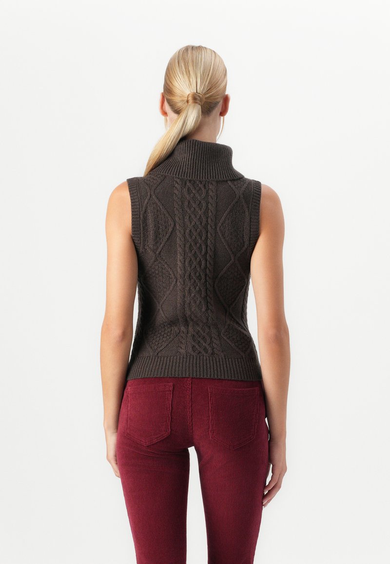 Femme avec une queue de cheval blonde portant un pull à col roulé sans manches en tricot marron et un pantalon en velours côtelé rouge, vue de dos.