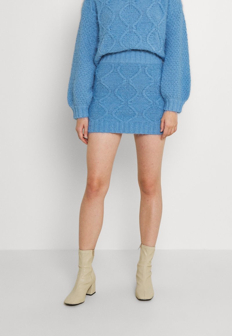 Stickad klädsel i ljusblått med en kortad tröja med diamantrand och en matchande figurnära minikjol. Beiga klackboots med blockhäll.