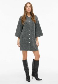 Pilka vilnos mišinio suknelė su apykakle, mygtukų priekyje ir trijų ketvirčių rankovėmis; derinama su juodais aukštakulniais batais iki kelių.