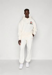 Felpa con cappuccio color crema con logo frontale, cappuccio con coulisse e tasca a marsupio; pantaloni jogger crema abbinati con polsini elasticizzati e marchio discreto.