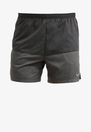 Homme en jogging portant un t-shirt noir, un short gris foncé et des chaussures de course Nike rouge vif sur un fond blanc uni.