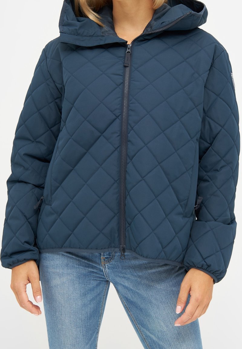 Femme portant une veste matelassée bleu foncé avec une fermeture éclair frontale et une capuche, associée à un jean bleu clair, vue du cou jusqu'aux hanches.