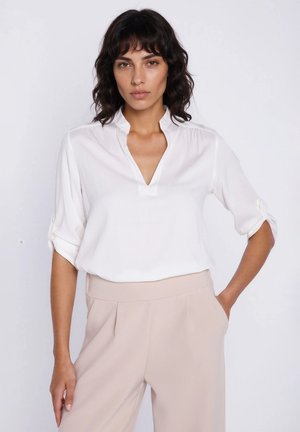 Kobieta o falujących ciemnych włosach, ubrana w białą bluzkę z dekoltem w kształcie litery V z podwiniętymi rękawami oraz jasnobeżowe, wysokiego stanu spodnie, stojąca na białym tle.