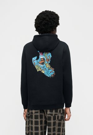 Tumejuuste ja tumedate lokkidega inimene kannab musta kapuutsiga pusa, millel on seljal värvikas abstraktne karjuva näo kujutis, koos mustade ja beežide ruuduliste pükstega.