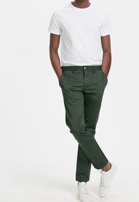 Gröna bomullsbukser med rak skärning, som har sidofickor och knäppning, tillsammans med en enkel vit t-shirt och vita sneakers.