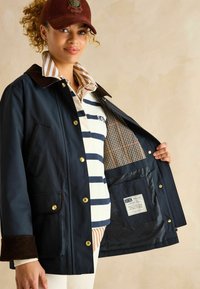 Jachetă impermeabilă de culoare albastru marin cu nasturi aurii, guler din catifea cord, căptușeală în carouri și două buzunare frontale. Purtați peste un tricou polo cu dungi.