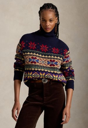 Femme aux cheveux tressés portant un pull à col roulé bleu marine avec des motifs géométriques colorés et un pantalon marron foncé avec une boucle de ceinture décorative.