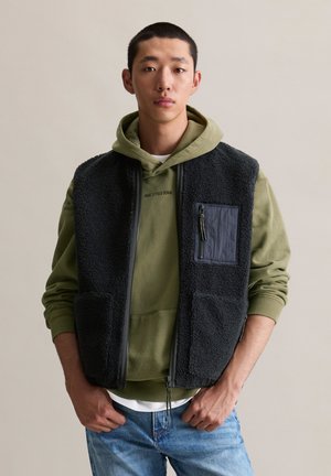 Black fleece vest with two front pockets and a zippered chest pocket, worn over a green hoodie and paired with blue jeans.