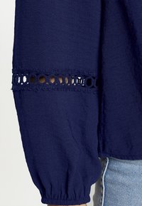 Blusa blu navy con maniche a palloncino; presenta dettagli in pizzo all'uncinetto ai polsini e un tessuto texture. Fits at the wrists.