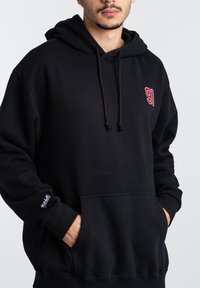 Schwarzer Kapuzenpullover aus weichem Stoff, mit einer Fronttasche, einstellbarem Kordelzug an der Kapuze und rotem, gesticktem "91" auf der Brust.