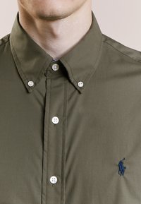 Olivgrünes Button-Up-Hemd mit spitzem Kragen, featuring weiße Knöpfe und ein kleines, dunkelblaues Logo, das auf der Brust gestickt ist. Glatte Stofftextur.