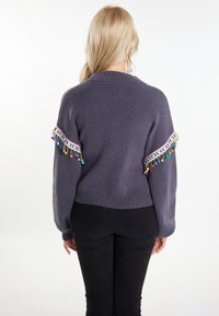 Lila kötött pulóver, színes pom-pom díszítéssel az ujjak mentén és bordázott szegéllyel, fekete skinny farmernadrággal párosítva.