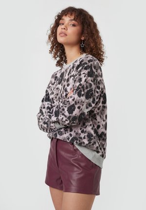 Kvinde med krøllet hår iført stjerneøreringe, en grå og sort leopardprint sweater og bordeaux lædershorts med armene over kors.