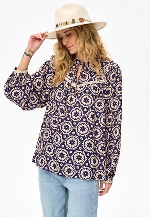 Vrouw draagt een blauw met beige blouse met patroon en kanten rand, lichtblauwe spijkerbroek en een beige hoed met brede rand, waarbij ze met één hand de rand van de hoed vasthoudt.