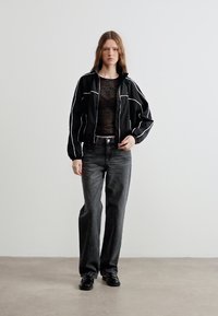 Νεαρή γυναίκα που στέκεται φορώντας μαύρο αθλητικό μπουφάν με λευκές ρίγες, διαφανές μαύρο τοπ, σκούρα γκρι τζιν και μαύρα παπούτσια, μπροστά σε απλό λευκό φόντο.