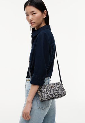 Young woman with straight black hair wears a navy shirt, light blue jeans, and a patterned crossbody bag with black strap over her shoulder.