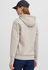 Hoodie beige avec capuche à cordon, présentant une texture douce et des poignets côtelés. Porté avec une casquette navy, vu de dos.