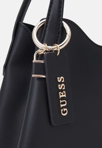 Détail d’un sac à main en cuir noir avec anneau doré et étiquette noire attachée présentant les lettres du logo GUESS en relief doré.