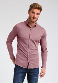 Langärmliges, knöpfbares Hemd aus sanftem hellrosa Stoff. Verfügt über einen klassischen Kragen, schmalen Schnitt und verdeckte Knopfblende. Kombiniert mit blauen Jeans.