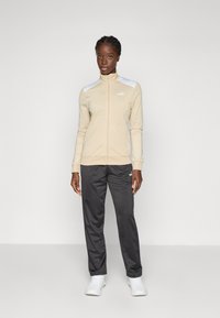 Beige athletic jacket with zip, white accents on shoulders, paired with dark gray pants and white sneakers. Smooth fabric, fitted design.