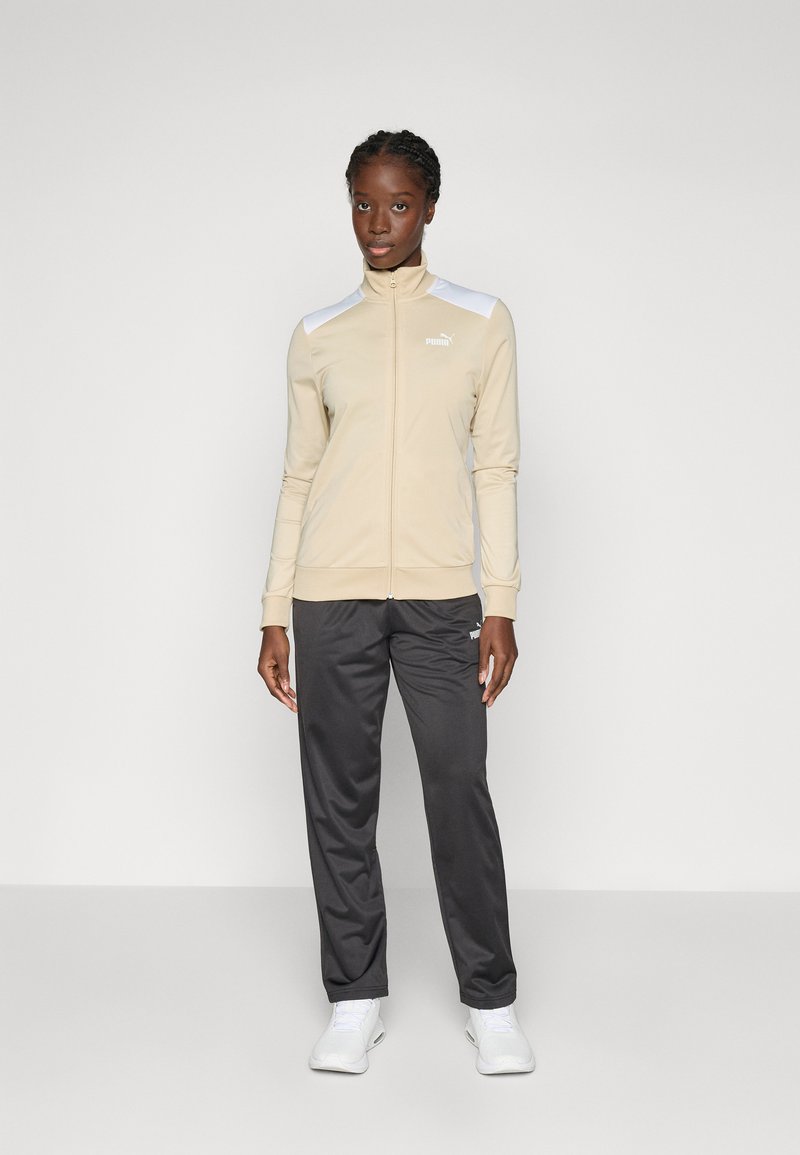 Beige athletic jacket with zip, white accents on shoulders, paired with dark gray pants and white sneakers. Smooth fabric, fitted design.