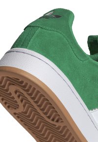 Gröna mockaskor med vit gummimellansula och brun strukturerad yttersula. Svart logotypdetalj på baksidan. Har en rundad tådesign.