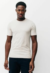 Gaiši bēša pieguļoša T-krekls ar īsām piedurknēm un apaļu kakla izgriezumu, izgatavots no gludas auduma. Valdā ar tumšām biksēm, izceļot vienkāršu dizainu.