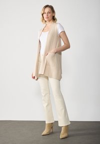 Beige ärmlös cardigan med öppen front och två fickor, bärs över en vit t-shirt och kombineras med krämfärgade byxor och beige högklackade skor.