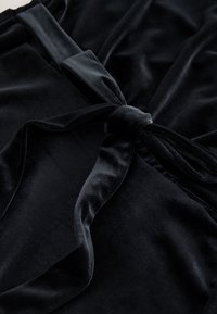 Tessuto di velluto nero con una trama liscia. Presenta un dettaglio con un nodo al centro e pieghe fluenti, che ne enfatizzano l'aspetto lussuoso.