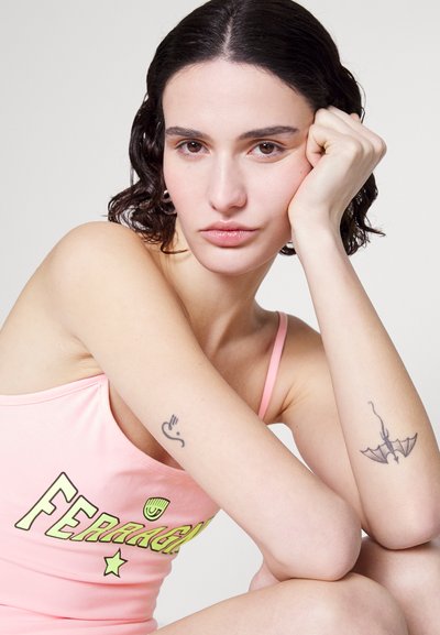 Ροζ αμάνικο τοπάκι με το κείμενο "FERRAGAMO", διαθέτοντας λείο υφές και στρογγυλή λαιμόκοψη. Ορατά τατουάζ σε κάθε χέρι. Ουδέτερο φόντο.