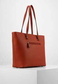 Sac fourre-tout en cuir rouge avec deux longues poignées, une poche avant zippée et des accents en métal doré, présentant un extérieur texturé.