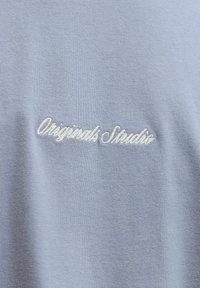 Hele sine sinine kangas pehme tekstuuriga, millel on valge tikkimisega tekst "Originals Studio." Lihtne disain, lisadeta.