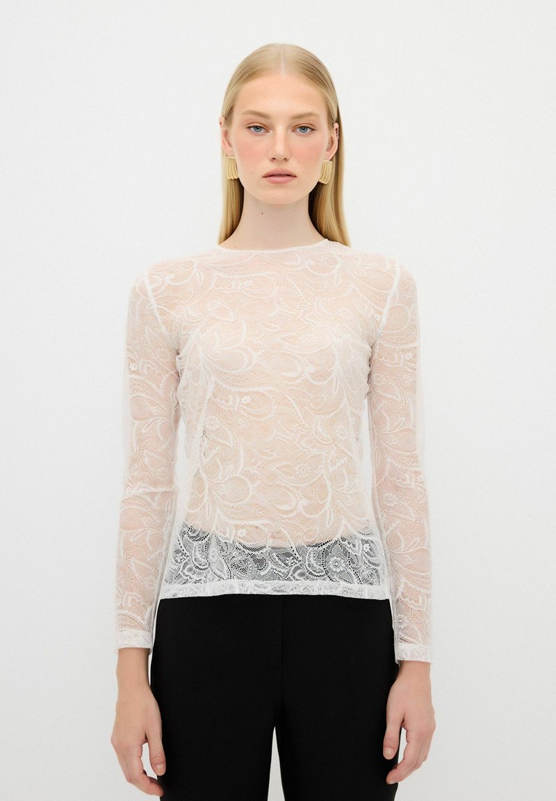 Top in pizzo a maniche lunghe in bianco trasparente, con motivo floreale e scollatura tonda. Abbinato a pantaloni neri a vita alta per un contrasto.