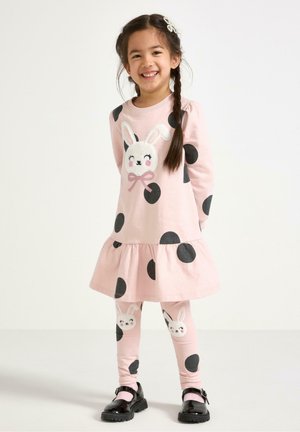 Lyserød kjole med sorte prikker, med et kaninansigt design; lagdelt nederdel og matchende leggings med kanindetaljer; parret med sorte sko.