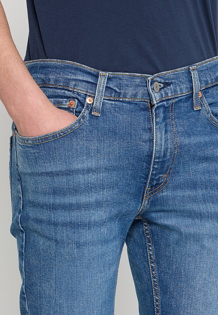 Hombre vistiendo jeans azules y una camisa azul marino, con la mano izquierda de manera casual en el bolsillo frontal, mostrando la textura del denim y los detalles de la costura.