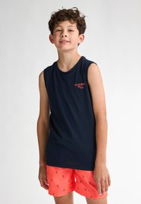 Granatowy bezrękawnik z małym czerwonym logo na piersi, w połączeniu z jasnymi koralowymi szortami z czarnym wzorem małych ptaków.