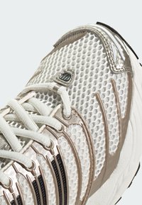 Biały siateczkowy sneaker z metalicznymi złotymi akcentami, teksturowaną cholewką, czarnymi paskami oraz sznurowadłami. Posiada wyściełany kołnierz i gumową podeszwę.