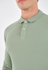 Lichtgroen, langmouw poloshirt van zachte katoen. Heeft een kraag en een twee-knopen placket met subtiele stikdetails.