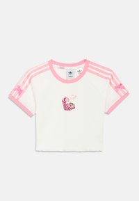 Fehér, felülvágott póló rózsaszín díszítésekkel, amelyen egy Cheshire macska grafika és rózsaszín Adidas csíkok találhatók a vállakon. Pamut anyagból.