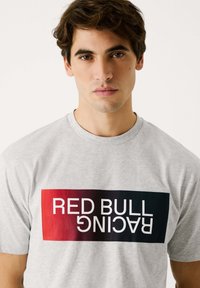 Pelēks kokvilnas t-krekls ar drosmīgu melnu un sarkanu gradienta taisnstūra grafiku, kurā ir teksts "RED BULL RACING" baltā krāsā.