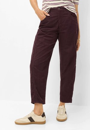 Frau trägt dunkelviolette weite Hose, gestreiftes Hemd eingesteckt und beige Sneakers mit dunkelbraunem Streifen, steht mit einer Hand in der Tasche.