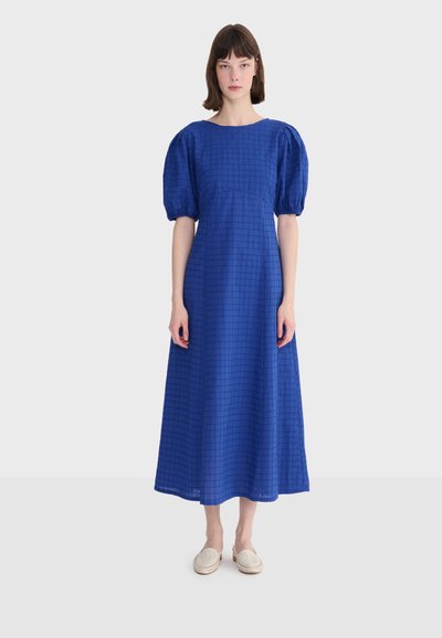 Femme portant une longue robe bleue avec des manches courtes bouffantes et un motif à carreaux subtil, debout les mains détendues le long du corps.