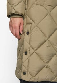 Close-up of a hand next to a beige quilted jacket with black snap buttons and diamond-pattern stitching on a white background.