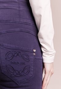 Lila denimoveraller med intrikata broderier på bakfickan, innehållande logotypdetaljer och dekorativa knappar.