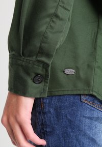 Manica di giacca di jeans verde con chiusura a bottone e patch logo metallico. Il tessuto è texturizzato con una finitura resistente.