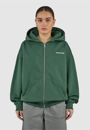 Młoda osoba z zaczesanymi do tyłu włosami, ubrana w leśnozieloną bluzę na zamek z kieszeniami z przodu oraz szare spodnie, na jednolitym tle.